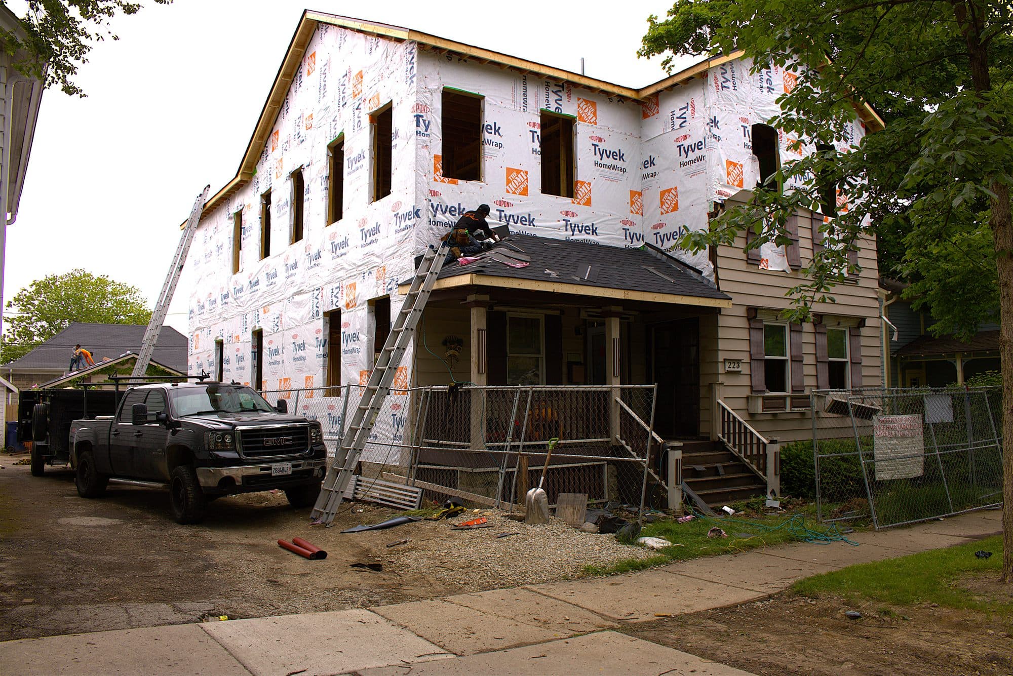 A black Revere One pickup and dump trailer parked at a Chicagoland home mid-restoration, its upper storey wrapped in housewrap and a crew member working on the porch roof