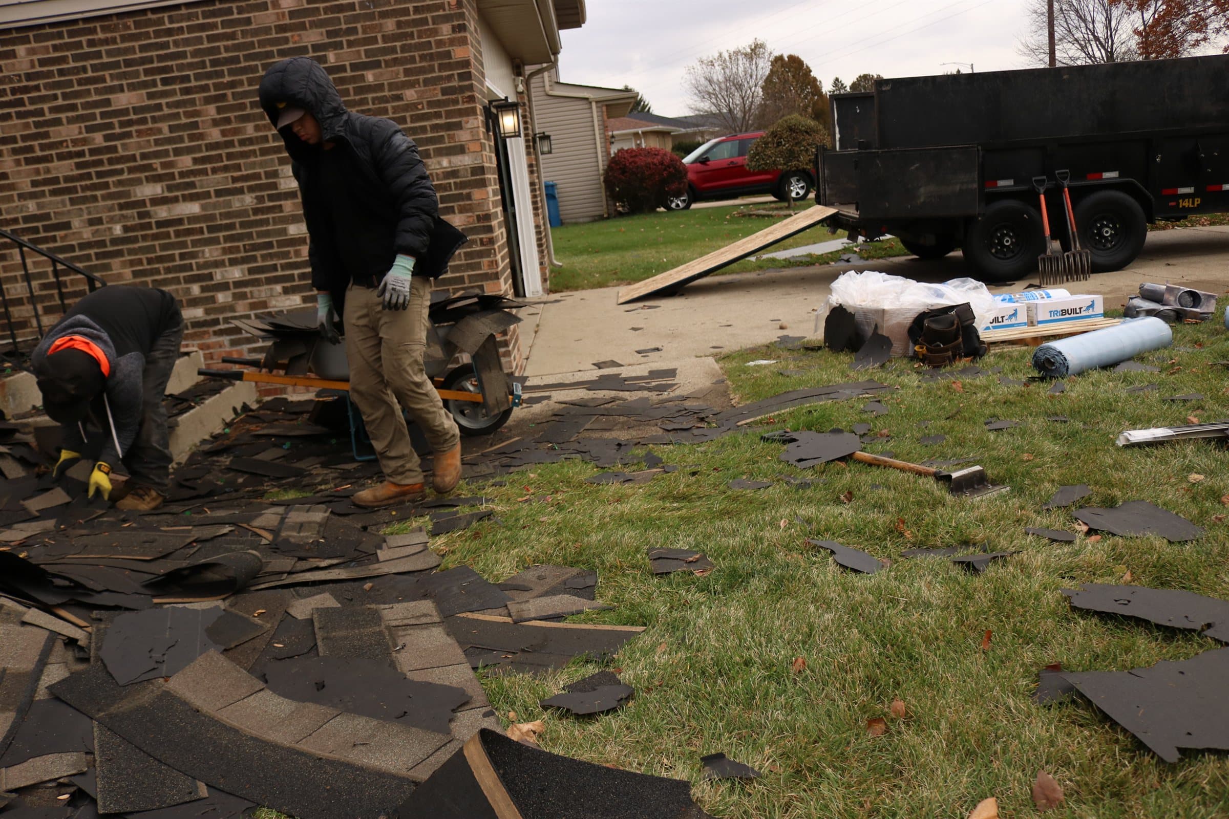 Stripped shingles covering a lawn beside a house, with crew clearing debris