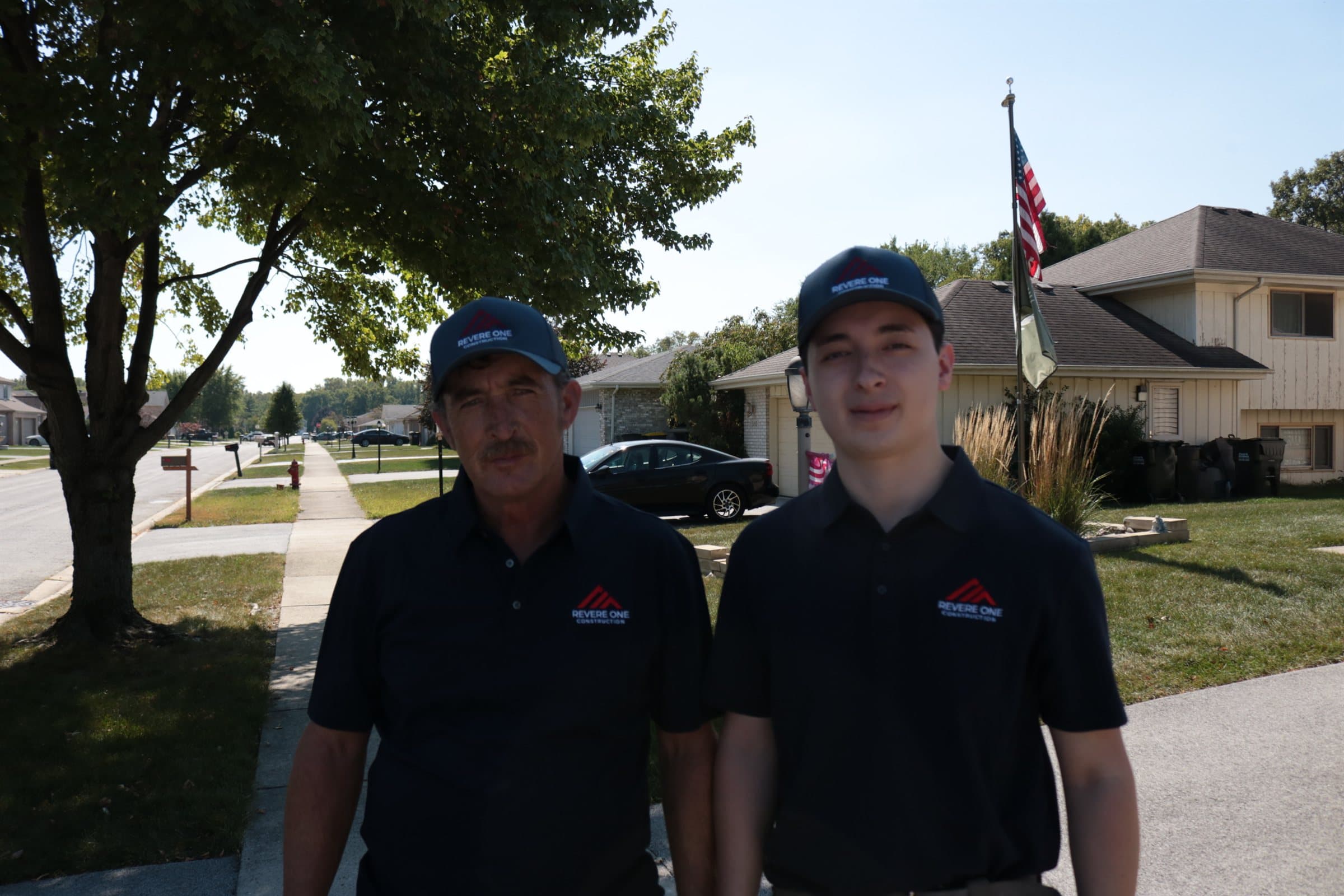 Two Revere One Construction team members in branded polo shirts and caps standing on a residential street