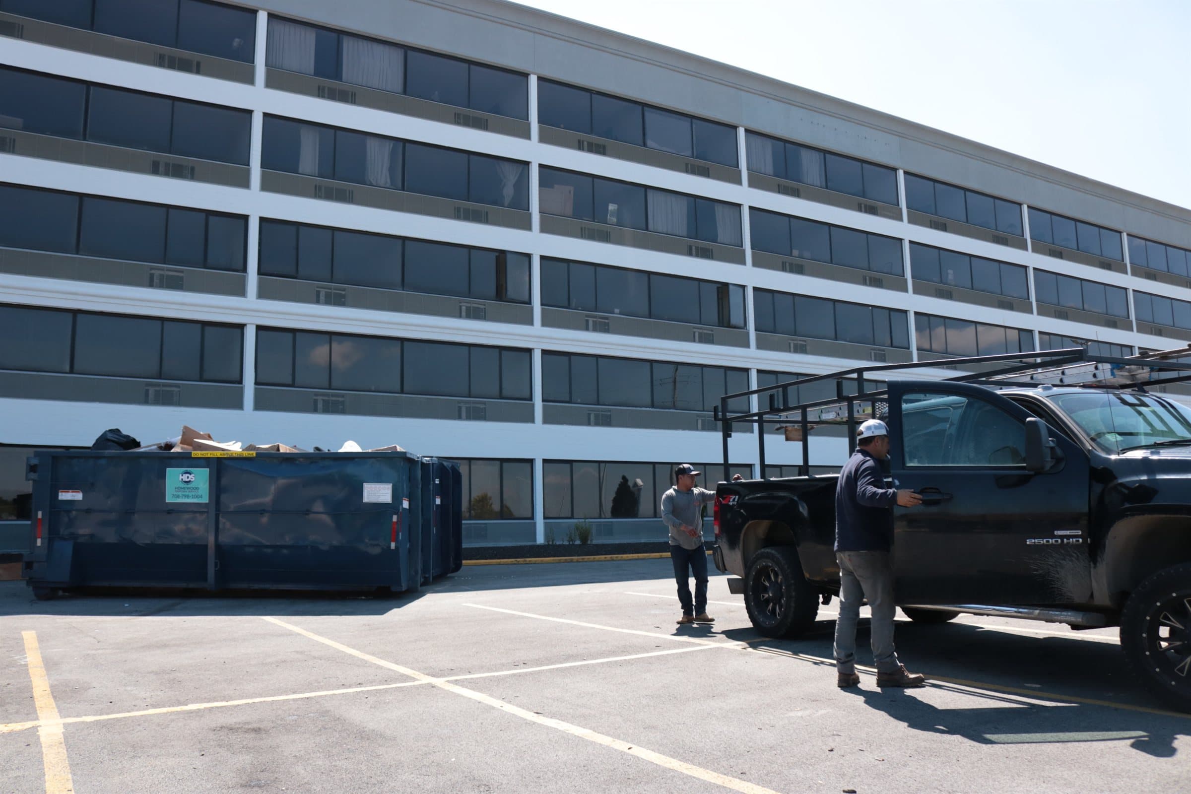 A Revere One pickup with a ladder rack and a debris container beside a multi-storey commercial building