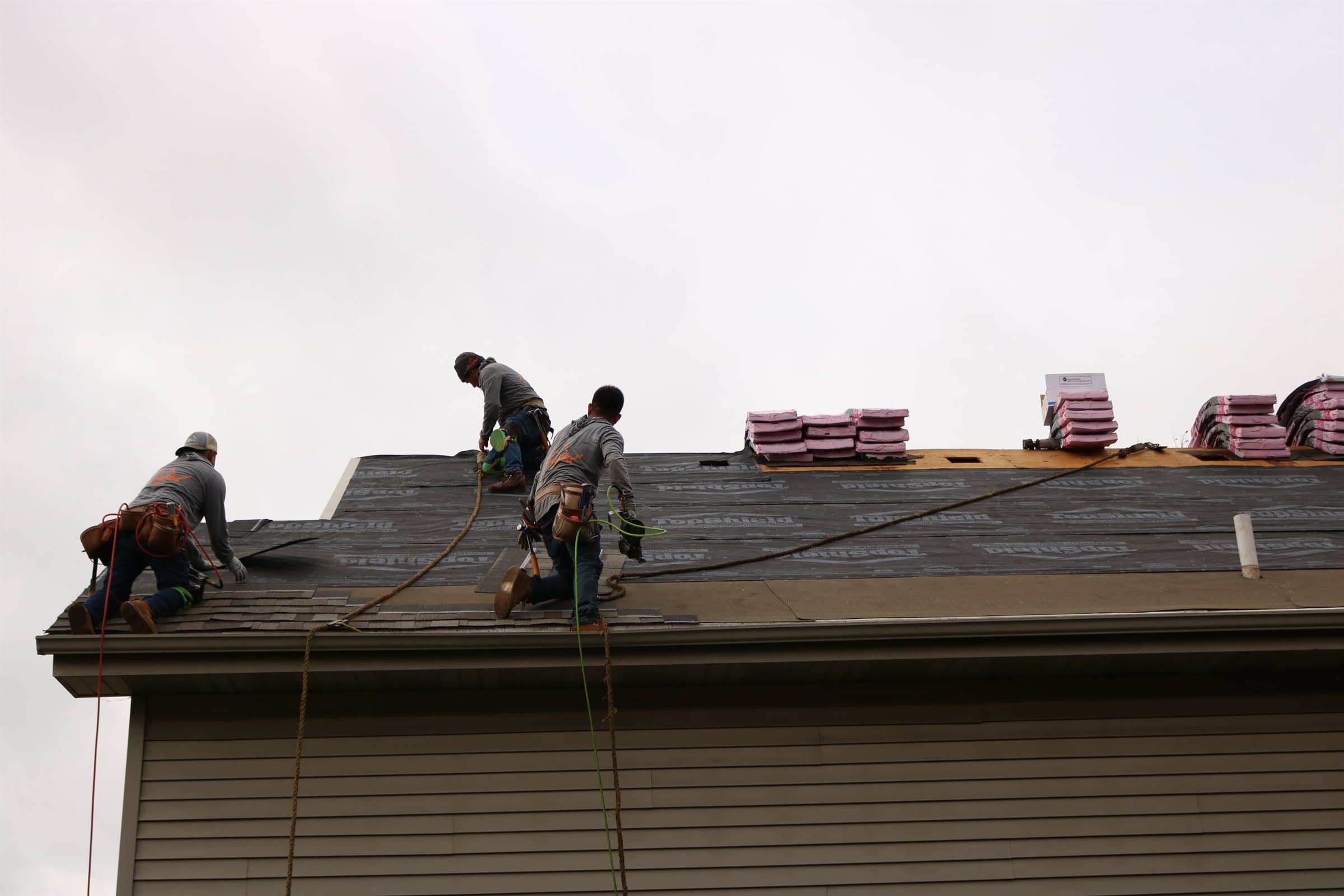 Three roofers working along a roof ridge against an overcast sky, wearing fall-arrest lines