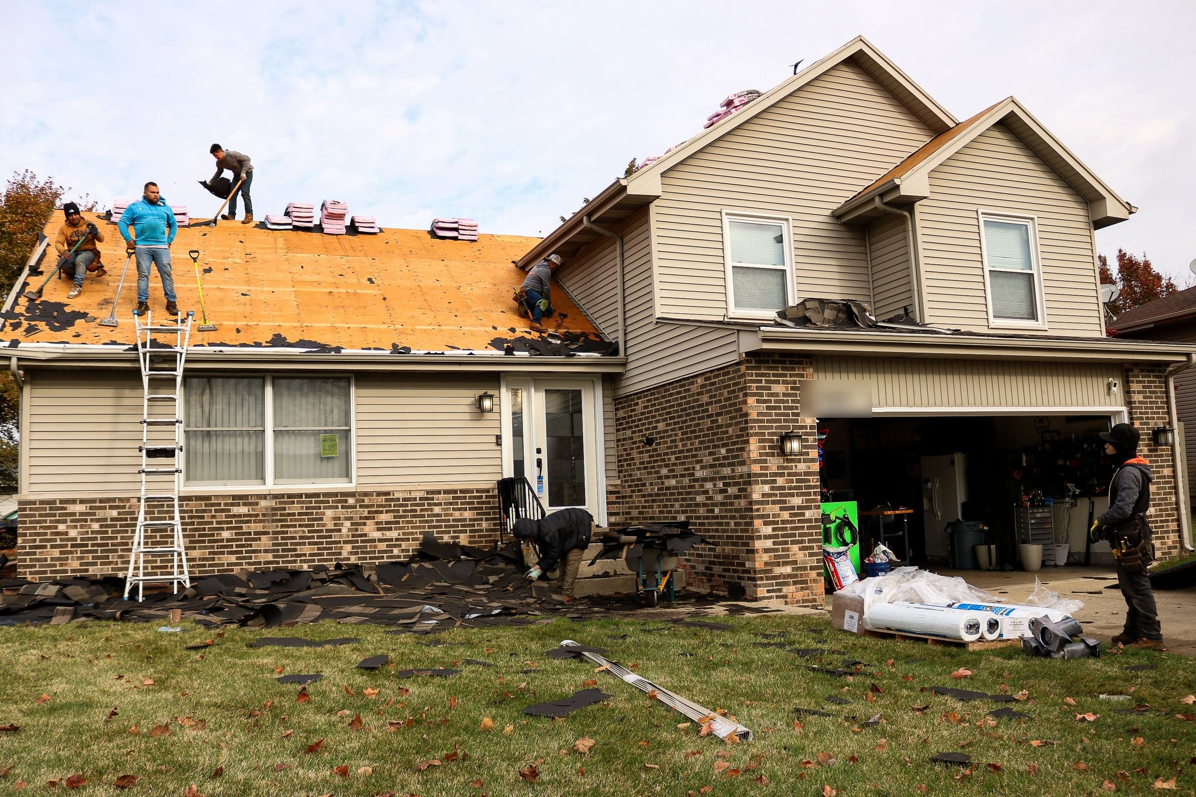 Roofers stripping the old roof to bare decking on a split-level home, with debris on the lawn below