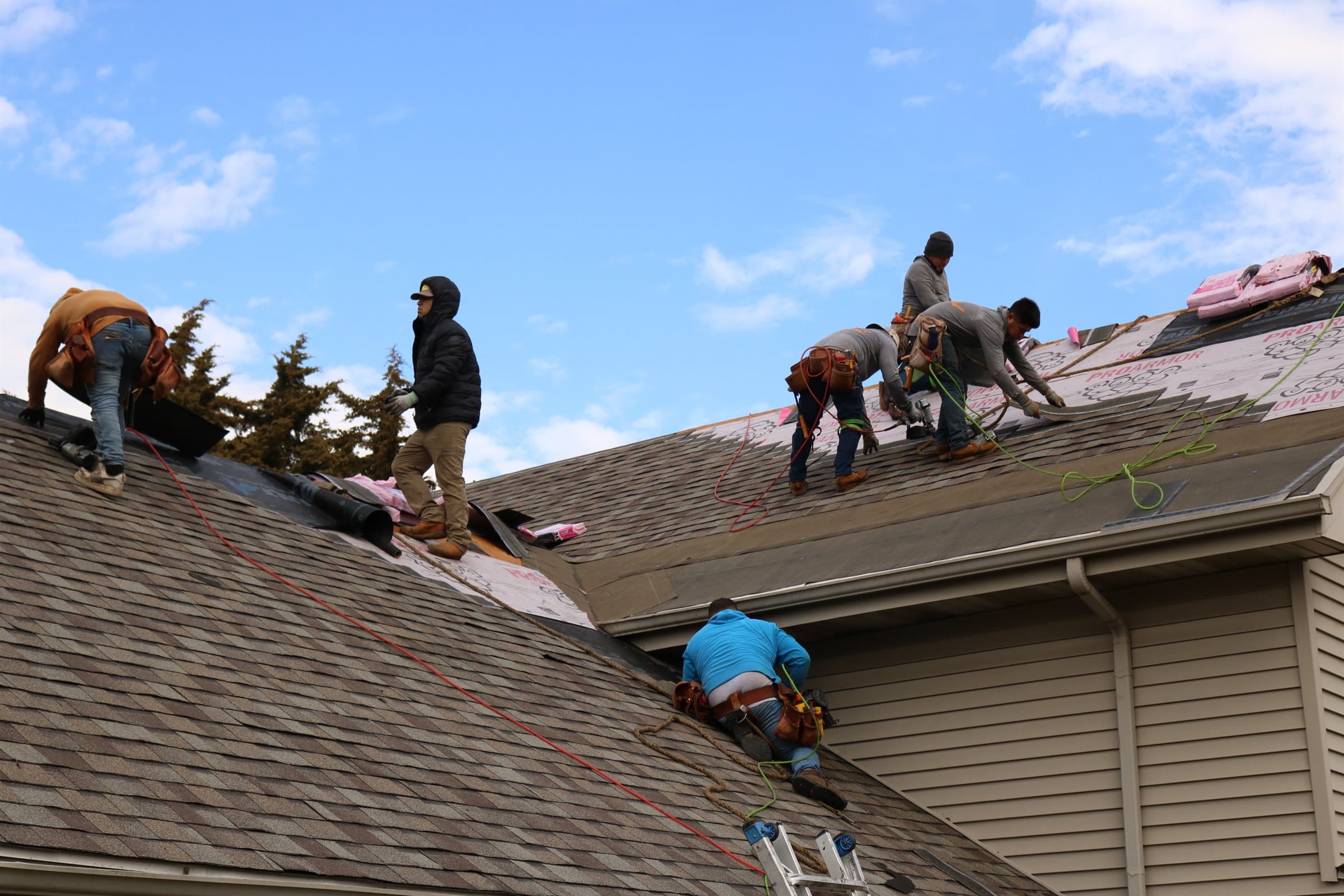 Roofers installing shingles across a second roof elevation under a blue sky