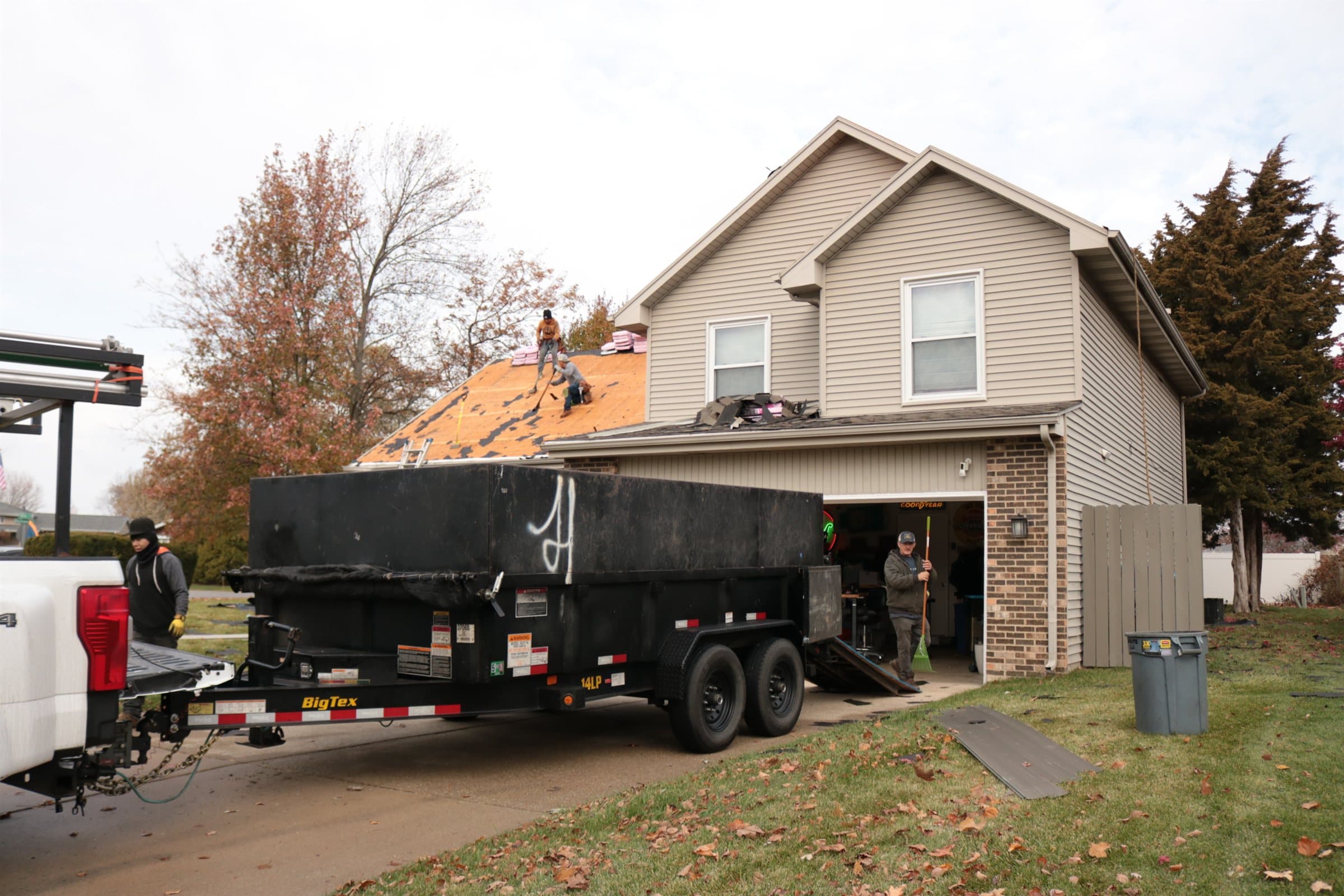 A dump trailer parked on a driveway beside a two-storey house with its roof being stripped