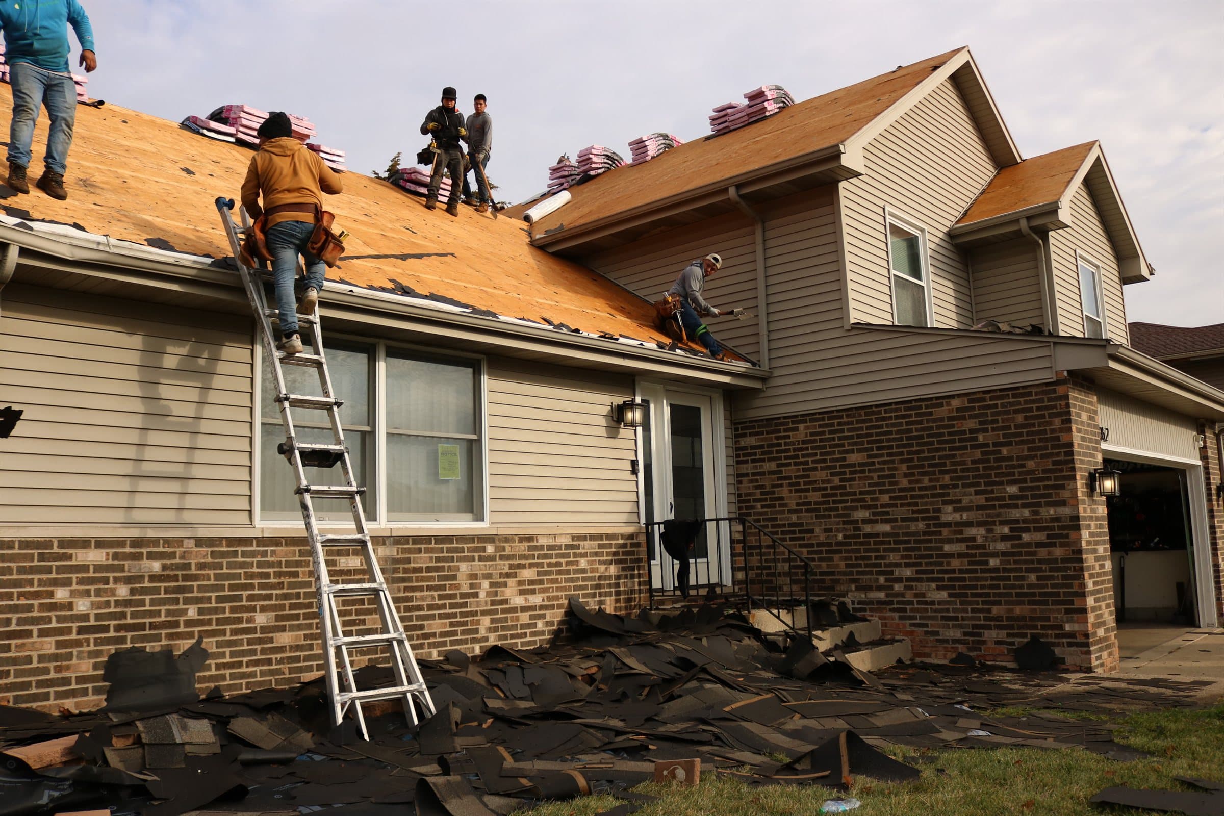 Stripped roof sections and shingle debris on the ground beside a brick and siding house