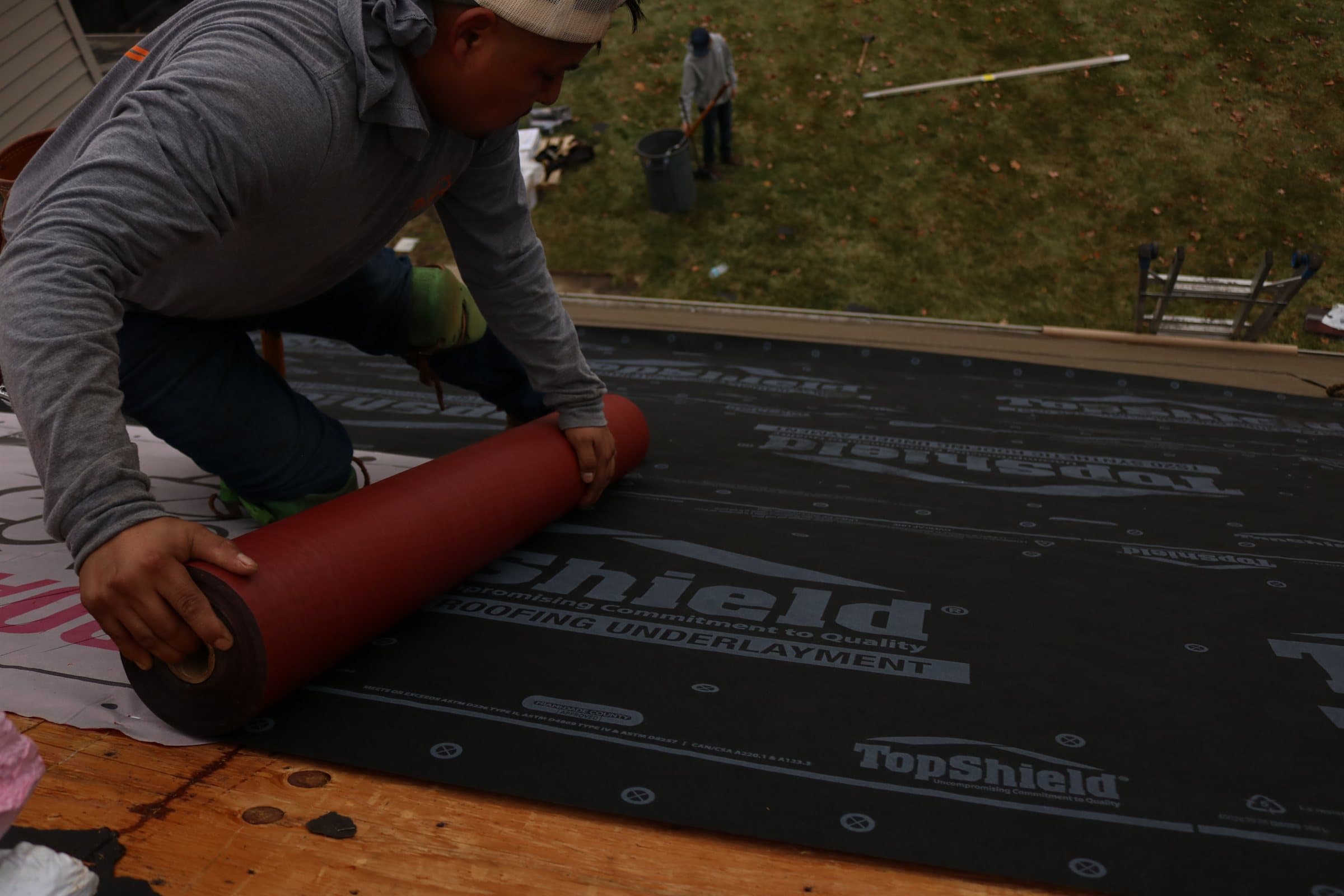A roofer unrolling a red membrane across black synthetic roofing underlayment