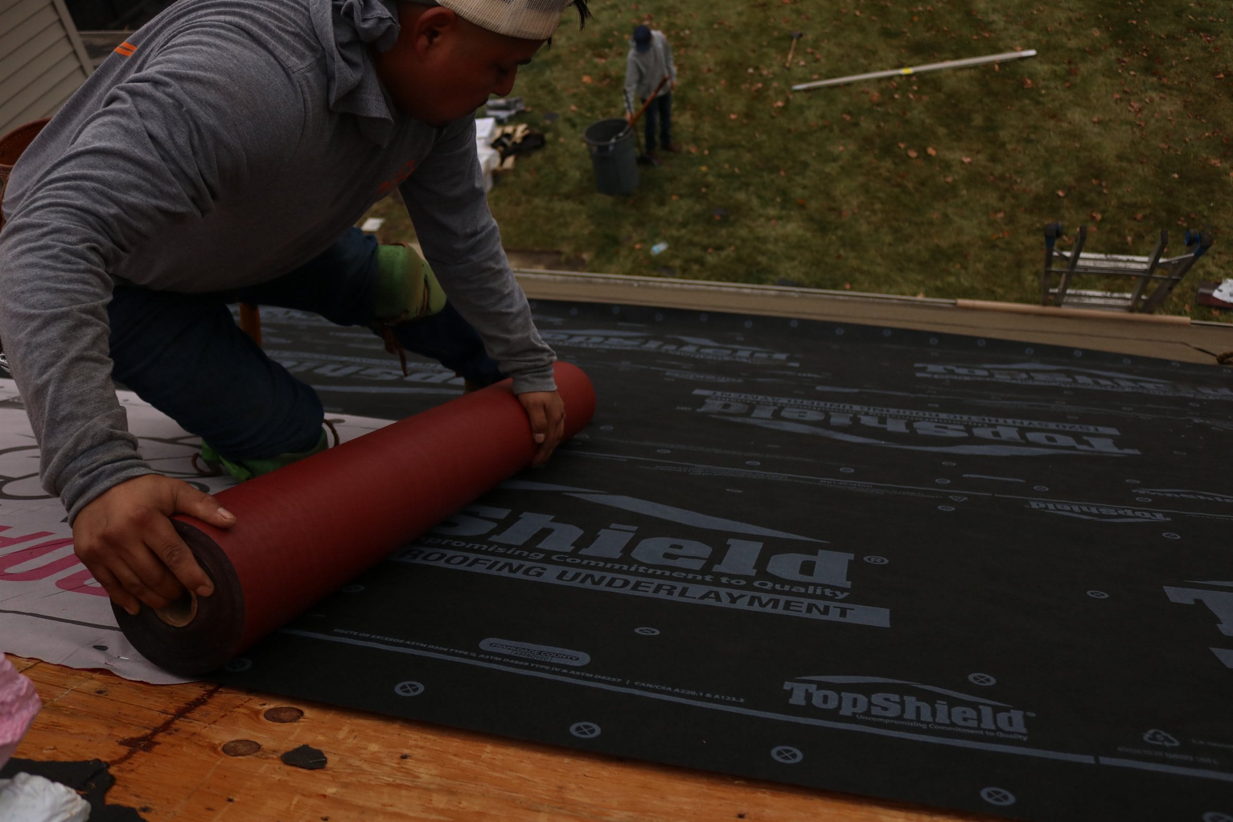A roofer unrolling a red membrane across black synthetic roofing underlayment